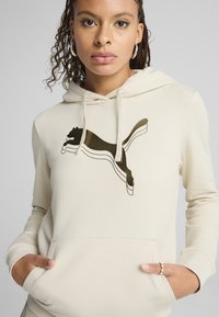 Bežová mikina s klokankou, na ktorej sa nachádza hnedý logotyp PUMA. Tkanina vyzerá mäkko, má voľný strih a nastaviteľnú kapucňu so šnúrkou.