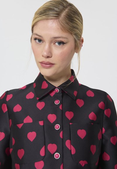 Schwarzes Hemd mit einem Muster aus roten Herzen, Kragen, vorderen Taschen und rosa Knöpfen mit dekorativen Akzenten. Glatte, satinartige Textur.