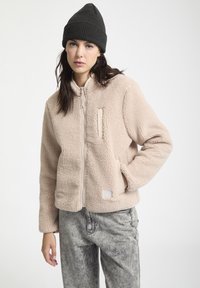 Beige fleece jack met rits aan de voorkant, staande kraag en twee zijzakken. Gecombineerd met grijze spijkerbroek en een zwarte gebreide beanie.