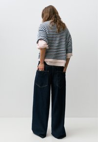 Person iført en stribet blå og hvid sweater med lyserøde manchetter og vidde mørke denimjeans, der står med hænderne i baglommerne.