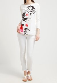 Top bianco a maniche lunghe con motivo floreale nero e rosa, abbinato a pantaloni bianchi attillati e sandali leggeri con dettagli dorati.