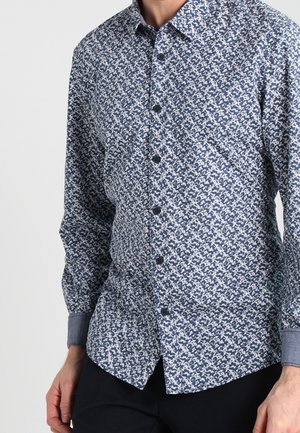 Homme portant une chemise à manches longues boutonnée avec un motif bleu et blanc complexe et un pantalon sombre.