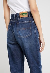 Mörkblå denimjeans med rak passform, har två bakfickor och en beige etikett med "TOMMY JEANS" broderat i grönt.