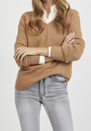 Person trägt beigen V-Ausschnitt-Pullover über weißem Hemd und hellgrauen Jeans, steht mit verschränkten Armen vor einfarbigem Hintergrund.