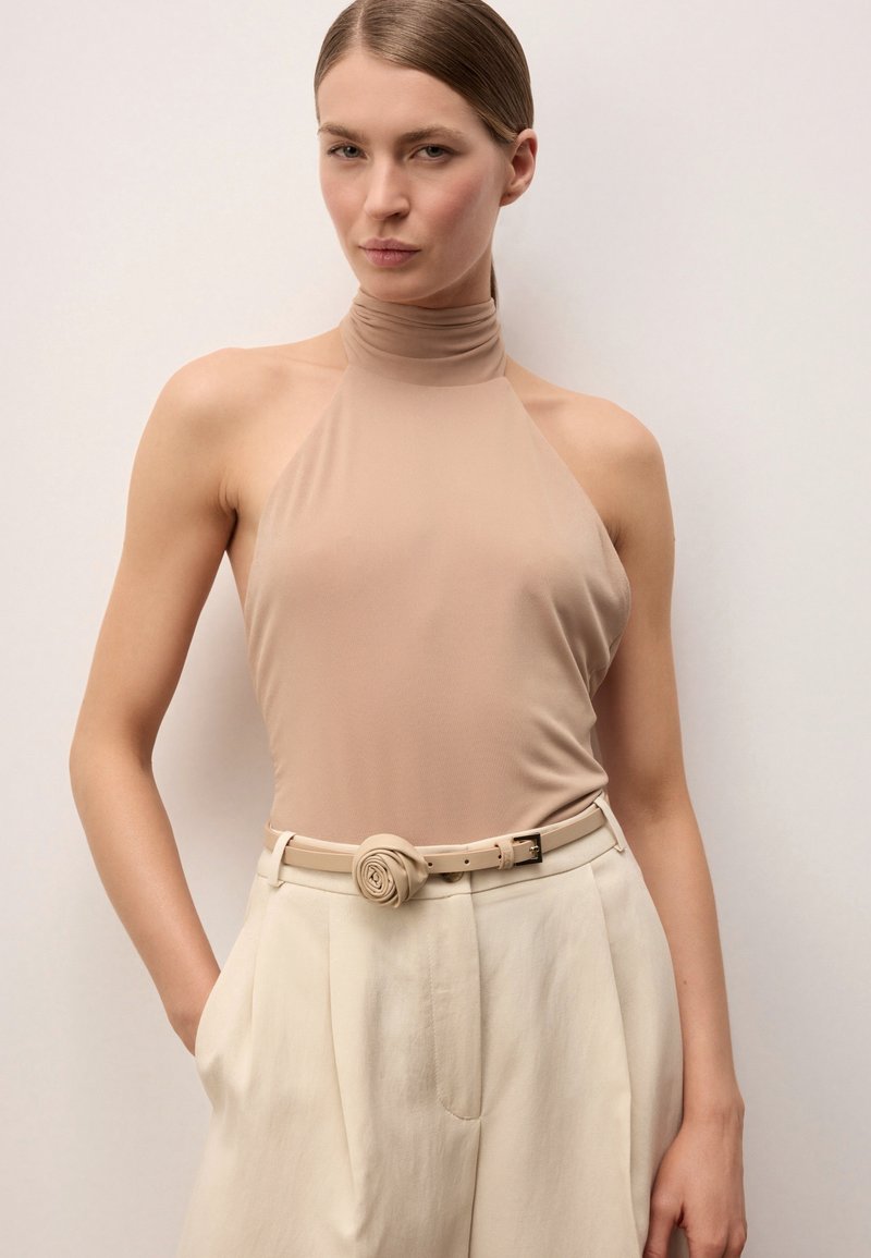 Žena v béžovém bezrukávovém halter topu a krémových kalhotách s vysokým pasem s růžovitě tvarovanou béžovou sponou, stojící před jednobarevným pozadím.