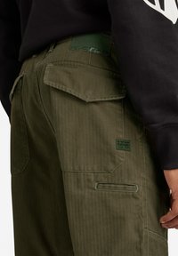 Olivgröna cargo-byxor i slitstarkt tyg. Funktioner inkluderar dubbla bakfickor, logotyp och subtilt herringbone-mönster.
