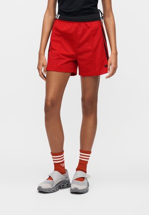 Persona con pantalones cortos deportivos rojos, calcetines a rayas rojas y blancas, zapatillas deportivas grises sin cordones y una camiseta negra con cordones laterales.