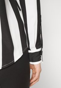 Schwarz-weiß gestreifte Bluse mit langen Ärmeln, glattem Stoff, geknöpften Ärmeln und geradem Saum. Nahaufnahme des Ärmels und der Hand.