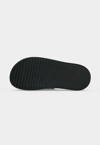 Semelle de chaussure noire avec des rainures horizontales pour l’adhérence, montrée à plat sur un fond gris clair.
