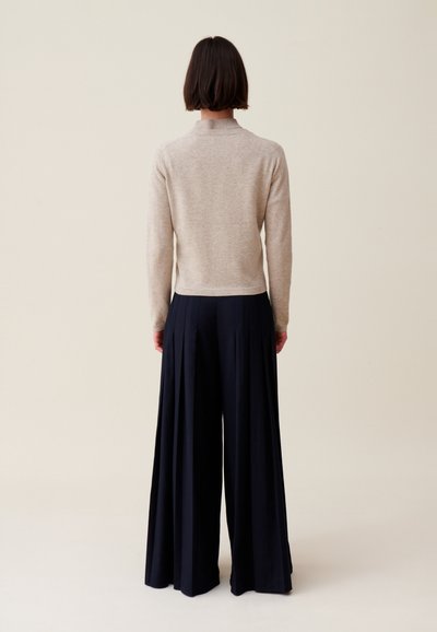 Pull en tricot beige à col montant, associé à un pantalon bleu marine taille haute, large et avec des plis.