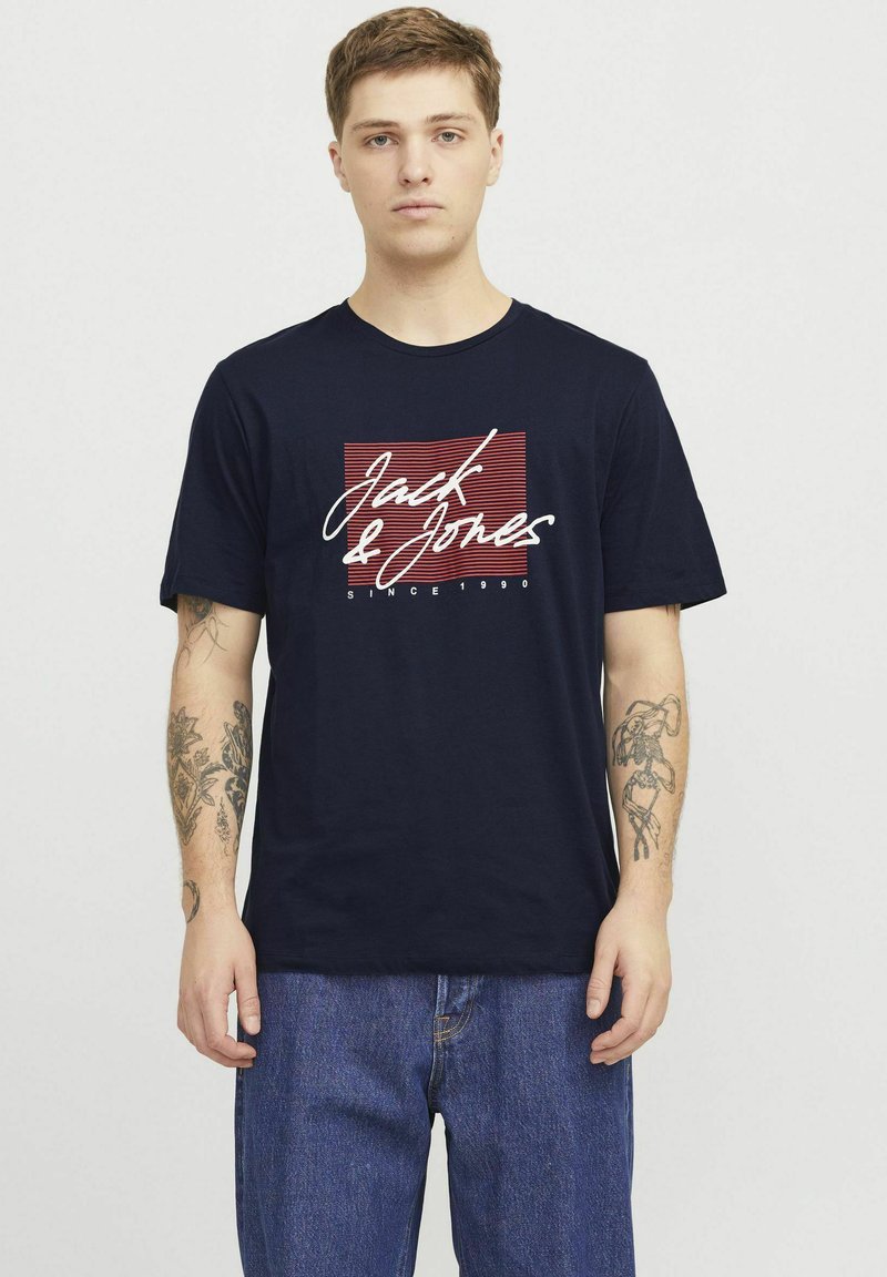 Granatowa bawełniana koszulka z białym napisem "Jack & Jones", czerwonymi poziomymi paskami oraz okrągłym dekoltem. Noszona z dżinsami.