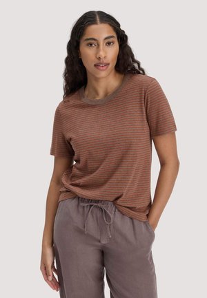 Frau mit langen dunklen Haaren, die ein kurzärmeliges gestreiftes Shirt in Rost- und Brauntönen und mauvefarbene Hose mit Kordelzug trägt, steht mit einer Hand in der Tasche.