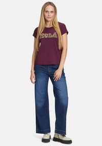 Maronfarbenes T-Shirt mit floralem "Hola"-Text, kurzen Ärmeln, kombiniert mit weiter geschnittenen blauen Jeans und beigen Stiefeln, vor einem schlichten Hintergrund.