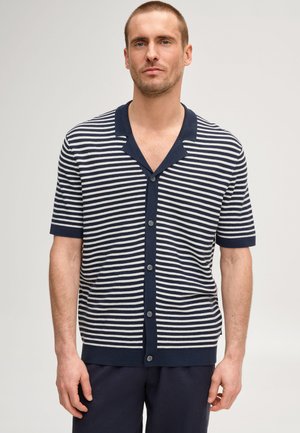 Uomo che indossa una camicia a maniche corte con righe orizzontali blu navy e bianche e colletto, in piedi davanti a uno sfondo uniforme.