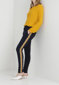 Blouse jaune à manches longues associée à un pantalon bleu marine avec des rayures jaunes sur les côtés. Escarpins blancs à enfiler avec un petit talon bloc.