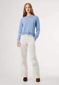 Svetlo modra pletena jopica s krožnim izrezom, v kombinaciji z slonokoščenimi širokimi hlačami in rjavimi čevlji s peto. Struktura se zdi mehka in sproščena.