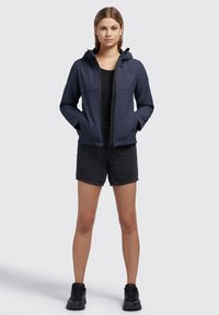 Marineblauwe waterdichte jas met een capuchon, rits aan de voorkant, twee borstzakken en elastische manchetten, gecombineerd met zwarte denim shorts en zwarte sneakers.