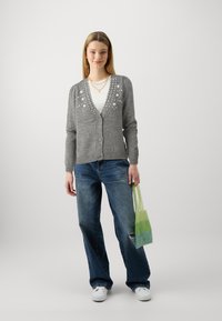 Cardigan grigio lavorato a maglia con decorazioni floreali, chiusura con bottoni e maniche lunghe. Abbinato a pantaloni di denim blu e una borsa a rete verde.