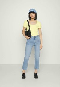 Top amarelo de manga curta, jeans retos azul claro, mala de ombro preta e sapatos slip-on pretos. Conjunto casual com um gorro azul.