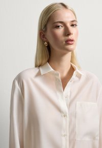 Blusa blanca de manga larga en tejido ligero con cuello puntiagudo, botonera frontal y un bolsillo de parche en el lado izquierdo del pecho.