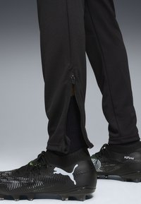 Pantaloni sportivi neri con zip alle caviglie e finitura testurizzata, abbinati a scarpe da calcio nere con un accento verde e logo bianco.