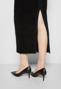 Robe midi noire à texture avec une fente sur le côté, associée à des talons aiguilles noirs à bout pointu avec un talon stiletto élancé.