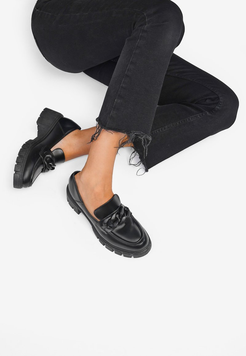 Zapatos negros de cuero tipo slip-on con suela gruesa, que cuentan con un detalle trenzado en la parte delantera, combinados con unos pantalones vaqueros negros deshilachados.