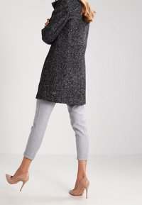 En kvinna med långt hår bär en mörk kappa, grå byxor och beigefärgade högklackade skor, stående mot en ljus bakgrund.