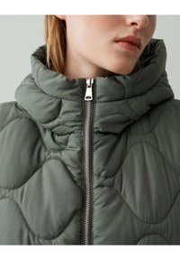 Die grüne gesteppte Jacke zeichnet sich durch einen hohen Kragen, einen senkrechten Reißverschluss und ein strukturiertes Muster aus weichem, glattem Stoff aus.