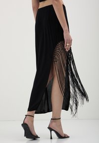 Gonna maxi nera con uno spacco alto, caratterizzata da dettagli frangiati intricati. Tessuto morbido con un design fluido. Abbinata a sandali neri con tacco.