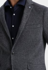 Uomo con una camicia button-up blu scuro indossata sotto un blazer grigio testurizzato con due bottoni e tasche frontali.