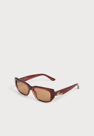 Lunettes de soleil rectangulaires teintées marron avec monture rouge foncé et logo doré "Guess" sur la branche, présentées sur une surface blanche.