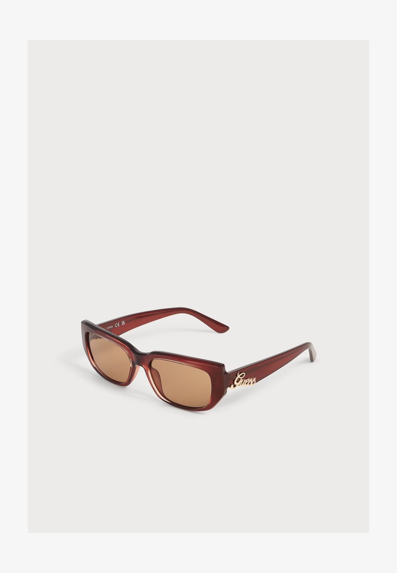 Lunettes de soleil rectangulaires teintées marron avec monture rouge foncé et logo doré "Guess" sur la branche, présentées sur une surface blanche.