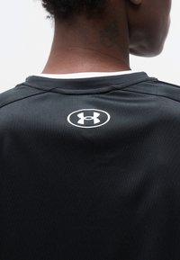 Κοντινό πλάνο στο άνω μέρος της πλάτης ενός ατόμου που φοράει μαύρο πουκάμισο με λευκό λογότυπο Under Armour κάτω από τον λαιμό και με ορατό τατουάζ στον λαιμό.