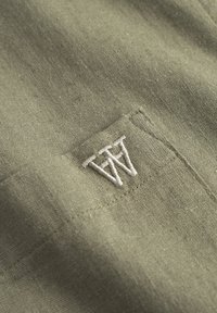 Olivovo zelená tkanina s textúrovaným povrchom; obsahuje vyšívané logo v bielej farbe. Pohľad zblízka, ktorý zvýrazňuje šitie a jemný materiál.