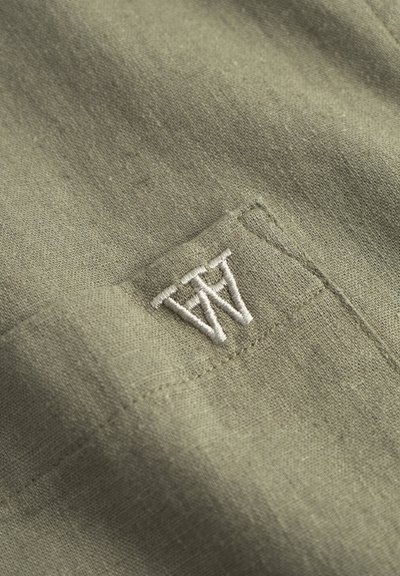 Olivovo zelená tkanina s textúrovaným povrchom; obsahuje vyšívané logo v bielej farbe. Pohľad zblízka, ktorý zvýrazňuje šitie a jemný materiál.