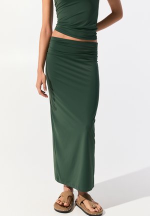 Longue jupe verte en tissu doux et extensible, avec une taille froncée et une finition lisse et soyeuse. Associée à des sandales brunes.