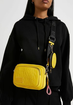 Żółta torebka crossbody z wytłoczonymi logo, z odpinaną okrągłą kieszonką na monety i regulowanym paskiem. Gładka tekstura i prostokątny kształt.