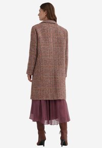 Kvinde iført en lang ternet frakke over en lilla lagdelt nederdel og brune hæle støvler, stående med ryggen til mod en hvid baggrund.