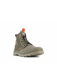 Bota alta de lona verde con suela resistente, tirador naranja y ojales, cordones elásticos negros, vista frontal en ángulo sobre fondo blanco.