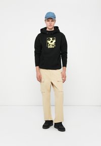 Black hoodie with a yellow skull graphic, beige cargo pants with side pockets, and black sneakers. Light blue cap completes the look.