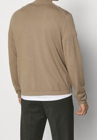 Uomo che indossa un maglione beige a maniche lunghe sopra una camicia bianca con pantaloni scuri, visto da dietro su uno sfondo chiaro e uniforme.