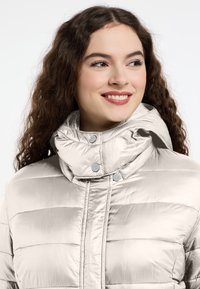 Jasnopłaszczowa beżowa kurtka puchowa z wysokim kołnierzem, z poziomym pikowanym wzorem i metalowymi zatrzaskami. Gładka, błyszcząca tkanina.