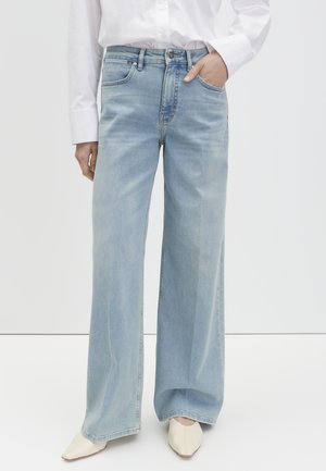 Persoon die lichtblauwe jeans met wijde pijpen draagt, witte puntige schoenen en een wit overhemd met knopen in de broek gestopt, met één hand in de zak.