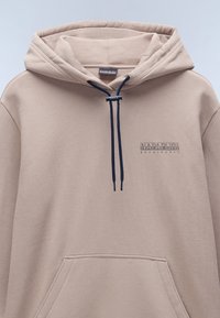 Sudadera con capucha beige fabricada en material suave. Presenta un bolsillo tipo canguro en la parte frontal y un cordón de color azul marino. Logo impreso en el pecho.