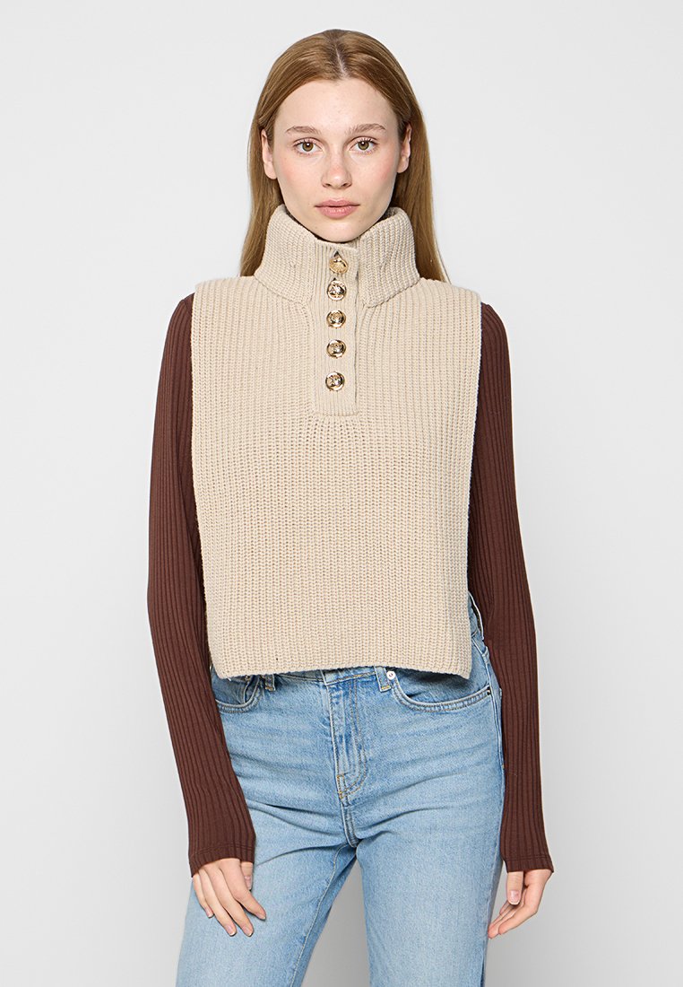 Beige gestrickter ärmelloser Pullover mit hohem Kragen, der mit fünf dekorativen Knöpfen verziert ist, über einem braunen Langarm-Shirt getragen.