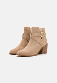 Bottines en daim beige avec un bout pointu, sangle ajustable avec boucle dorée et talon empilé. Texture lisse et design minimaliste.