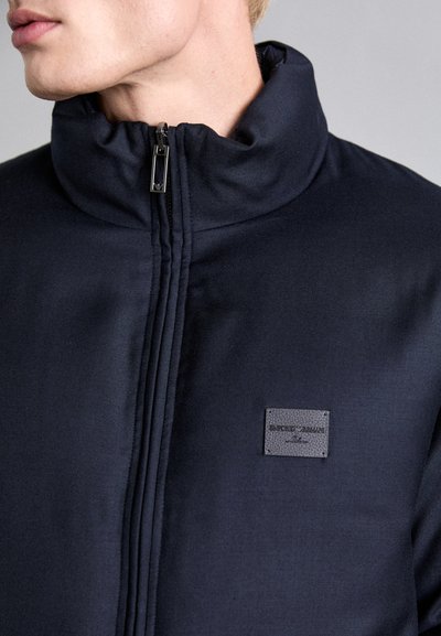 Navy blue jacket with a high collar, zip closure, and textured fabric. Features a branded black leather tag on the front.
