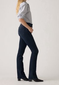 Femme en jean bootcut bleu foncé, bottes noires à bout pointu et chemise blanche à manches longues avec des rayures verticales noires, debout de profil.
