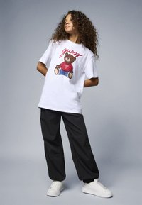 Vit t-shirt med en björngrafik i rött och blått, kombinerad med svarta vida byxor och vita sneakers, mot en grå bakgrund.